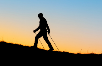Silhouette d'une personne pratiquant la marche nordique au coucher du soleil, avec des bâtons de marche sur un terrain en pente.